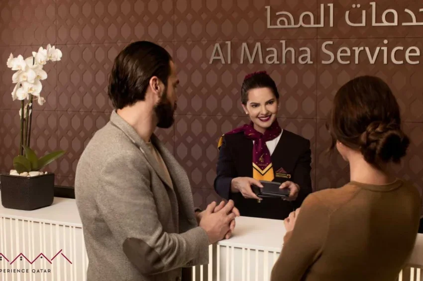 A man and woman stand at a reception desk, speaking with a smiling female staff member in uniform. The backdrop displays Al Maha Services in English and Arabic, highlighting their commitment to exceptional hospitality. A white orchid is on the counter.