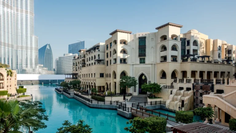 Modern beige buildings with arched windows line a turquoise canal, surrounded by lush greenery and elegant 4-star hotels in Qatar. Tall glass skyscrapers rise in the background under a clear blue sky.