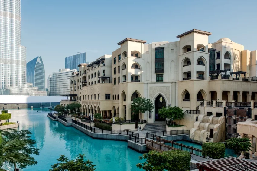 Modern beige buildings with arched windows line a turquoise canal, surrounded by lush greenery and elegant 4-star hotels in Qatar. Tall glass skyscrapers rise in the background under a clear blue sky.