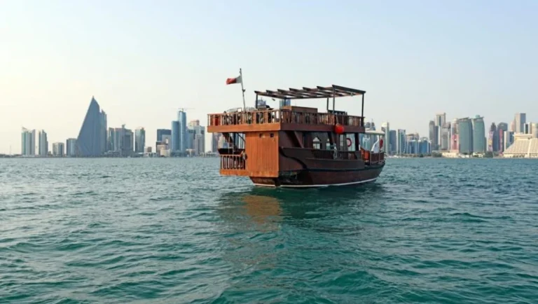A traditional wooden dhow boat floats on calm water with a modern city skyline in the background under a clear sky, capturing the essence of dhow boat rental in Doha.