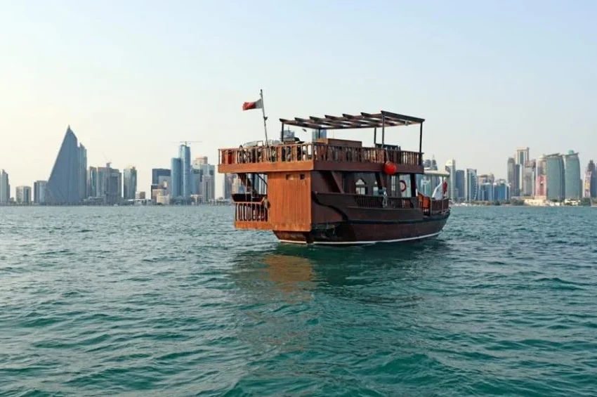 A traditional wooden dhow boat floats on calm water with a modern city skyline in the background under a clear sky, capturing the essence of dhow boat rental in Doha.