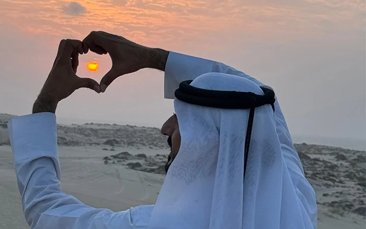 A man wearing traditional Middle Eastern attire forms a heart shape with his hands, framing the サンセット in a desert landscape under a partly cloudy sky—an iconic moment from a デザートサファリ or 半日ツアー adventure.