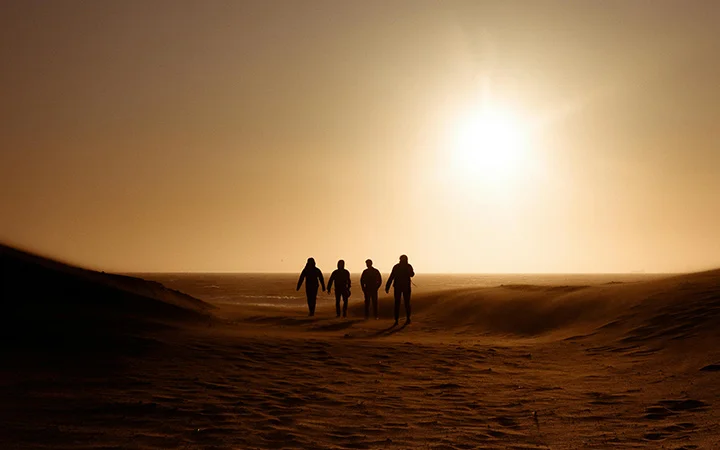 Four people walk together on a sandy beach at sunset, silhouetted against the bright sun and an orange sky, capturing the tranquility of a 半日ツアー.