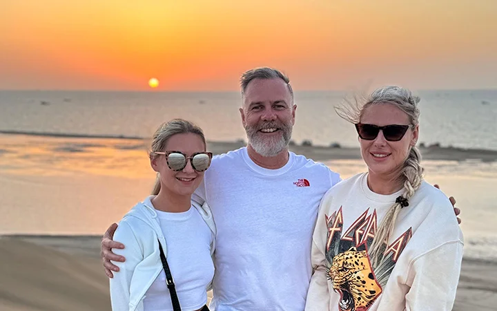 Three people stand smiling on a beach at sunset. The sun is low over the horizon, casting a warm glow—perfect for a サンライズ or デザートサファリ. The man in the center has gray hair and a beard; the two women wear sunglasses and light clothing.