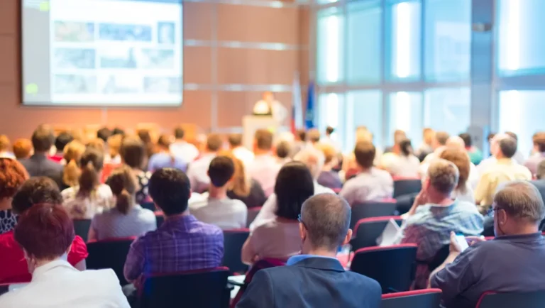 A large group of people sits in rows facing a speaker at the front of a bright conference room, watching a presentation projected on a screen, organized by a leading MICE DMC in Qatar.