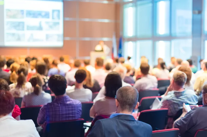A large group of people sits in rows facing a speaker at the front of a bright conference room, watching a presentation projected on a screen, organized by a leading MICE DMC in Qatar.