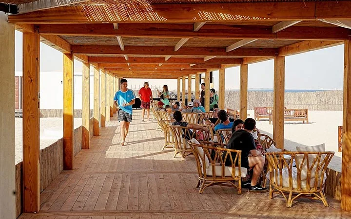 People sit and relax on bamboo chairs under a shaded wooden walkway on a sandy beach. Some chat while others look towards the sea, capturing the laid-back vibe of overnight camping by the shore as someone walks by carrying items.