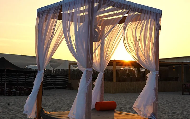 A wooden cabana draped with sheer white curtains stands on sandy ground at sunset, the sun casting a warm glow—an inviting spot for camping. Inside the cabana, a red cylindrical pillow adds comfort to this serene overnight camping retreat.