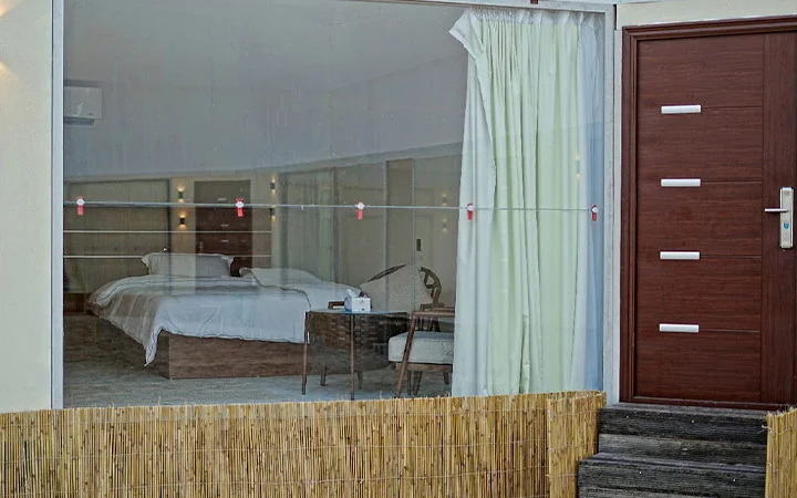 A hotel room with two beds seen through a large glass window, next to a wooden door and steps—perfect for those seeking the comfort of a modern interior after overnight camping. Curtain partially open, it reveals a tidy space with a small table and chair.