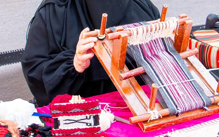 A person in a black garment weaves black, white, and red threads on a small wooden loom at a Qatari farm, with woven fabric pieces and tools spread out on a red table before them.
