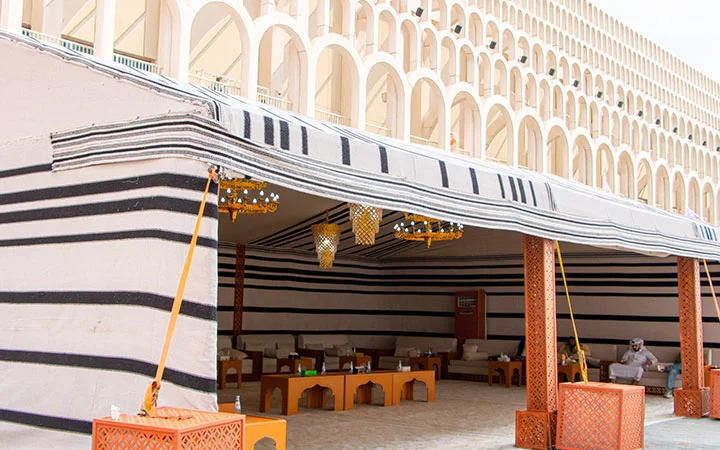 A traditional Arabic tent with black and white stripes stands in front of an arched building, evoking a Qatari Farm Experience. Inside are low wooden tables, cushioned seating, and decorative lanterns. A person sits on a sofa to the right.