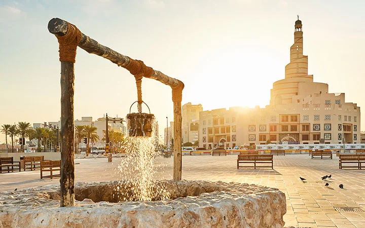 A stone well with a wooden frame and bucket pouring water in a sunlit square, with benches, palm trees, and the spiral minaret of Fanar Mosque in the background—a highlight on any ultimate guide to a Doha tour.