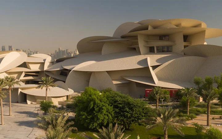 A modern building with curved, overlapping beige panels resembling desert rose crystals, surrounded by palm trees and greenery—a must-see on your Tour Doha adventure—stands out against a clear sky with a cityscape in the background.