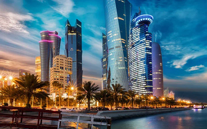 A vibrant cityscape at dusk with modern, uniquely shaped skyscrapers illuminated by lights—perfect for a Four Hours Tour Doha—palm trees line the busy waterfront road beneath a partly cloudy sky.