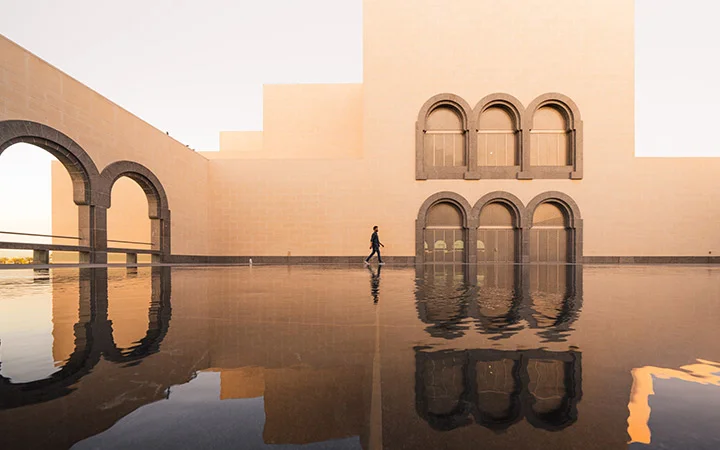 A person walks along the edge of a reflective pool in front of the modern beige National Museum of Qatar, its arched windows and doorways glowing softly under the evening light.