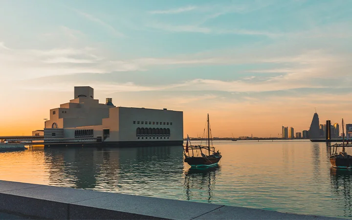 A modern, angular building sits by the water at sunset during a Doha Panoramic Tour, with traditional wooden boats floating nearby and the city skyline—including the National Museum of Qatar—visible in the distance under a colorful sky.
