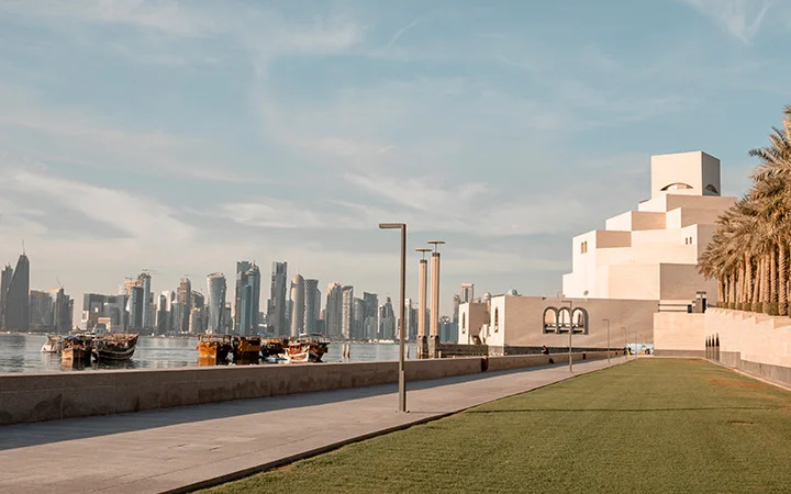 A waterfront view of Doha with modern skyscrapers, traditional boats on the water, a palm-lined pathway, and the striking white geometry of the National Museum of Qatar on the right—perfect scenery for a relaxing lunch or a Doha Panoramic Tour.