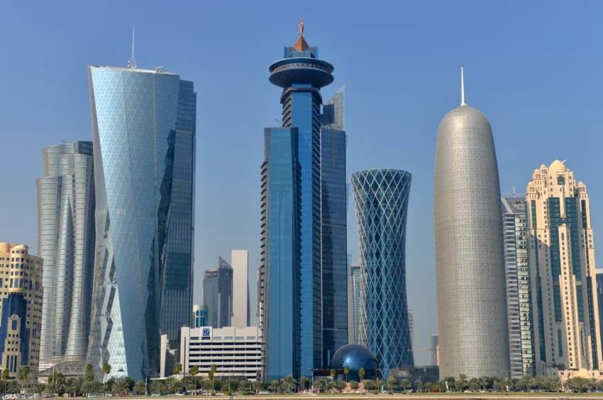 A city skyline with modern, uniquely shaped skyscrapers under a clear blue sky in Doha, Qatar. The mix of curved, cylindrical, and angular designs highlights some of the top architectural attractions in Doha.