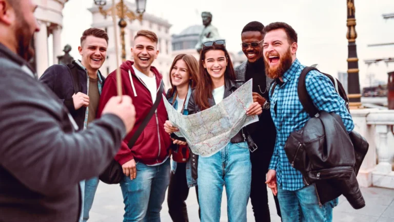A group of six young adults stand outdoors, smiling and laughing as one holds a map. They appear to be tourists exploring with Tour Guides in Qatar, with historic buildings and statues visible in the background.