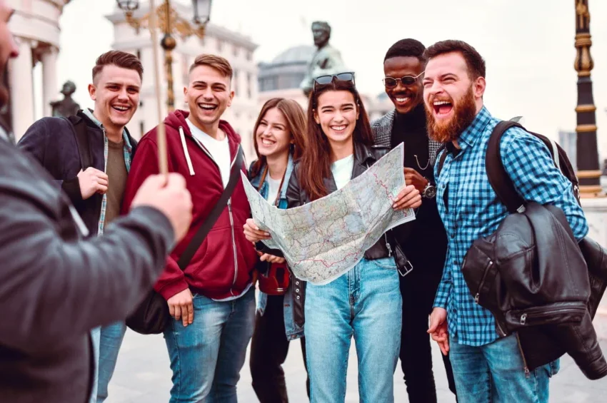 A group of six young adults stand outdoors, smiling and laughing as one holds a map. They appear to be tourists exploring with Tour Guides in Qatar, with historic buildings and statues visible in the background.