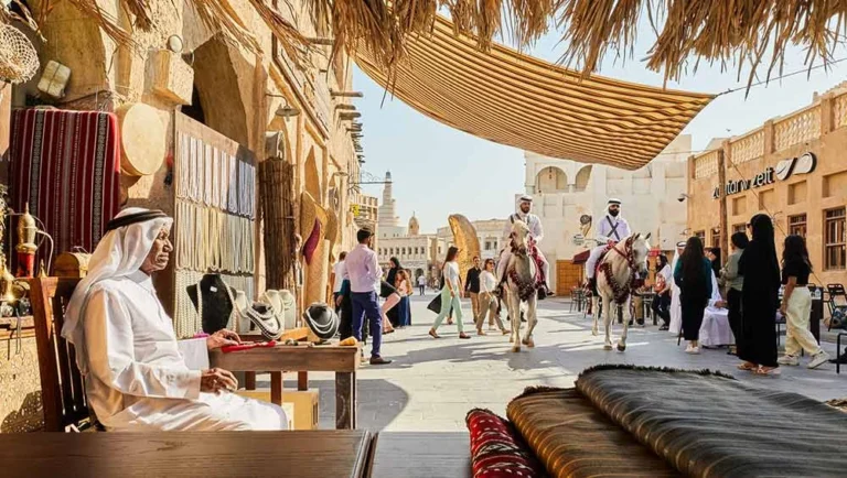 A traditional Middle Eastern market scene captures Qatar’s heritage, with a man in white attire at a stall of woven goods, people strolling past, and two men riding horses through the sunlit street.