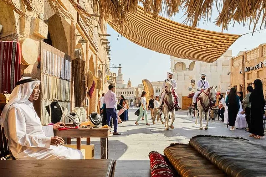 A traditional Middle Eastern market scene captures Qatar’s heritage, with a man in white attire at a stall of woven goods, people strolling past, and two men riding horses through the sunlit street.