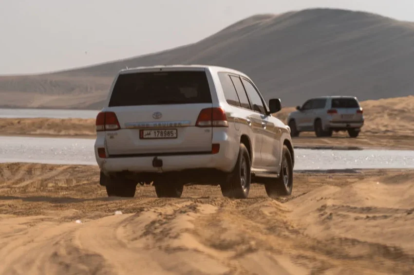 Two white SUVs drive on sandy terrain near water with large sand dunes in the background under a clear sky, capturing the thrill of a Desert Safari Qatar adventure.