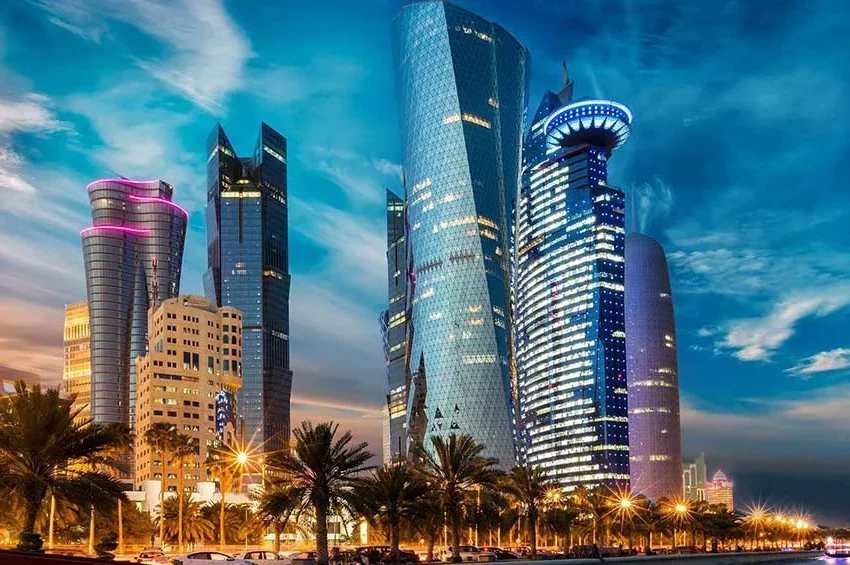 A cityscape at dusk shows modern, illuminated skyscrapers with unique glass facades, palm trees along vibrant streets, and streetlights glowing—capturing the lively atmosphere during Qatar Events under stunning Qatar weather.