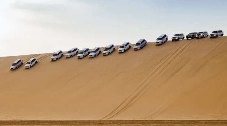A row of SUVs drives diagonally down a steep, sandy desert dune under a pale sky, their tire tracks marking the sand—an exhilarating scene from one of the Top 10 Unique Adventure Experiences in Qatar.