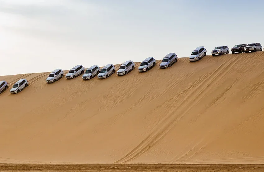 A row of SUVs drives diagonally down a steep, sandy desert dune under a pale sky, their tire tracks marking the sand—an exhilarating scene from one of the Top 10 Unique Adventure Experiences in Qatar.