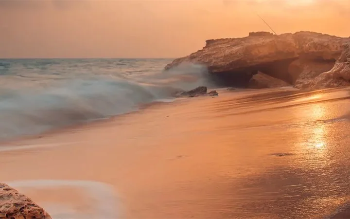 Golden sunlight reflects off the sandy shores of Fuwairit Beach, Qatar, as gentle waves wash ashore. Rocky formations sit near the water, and a soft orange sky suggests either sunrise or sunset.