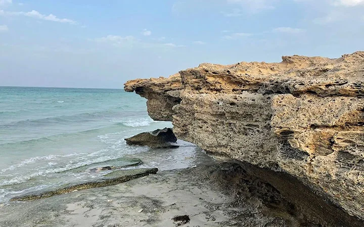 Large, jagged rock formation juts out over the sandy shores of Fuwairit Beach Qatar, framed by gentle blue-green waves and a partly cloudy sky.