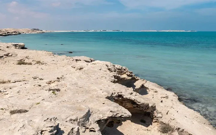 Rocky shoreline with light beige rocks meeting clear turquoise water under a partly cloudy sky at Fuwairit Beach Qatar, with a distant horizon visible across the calm sea.