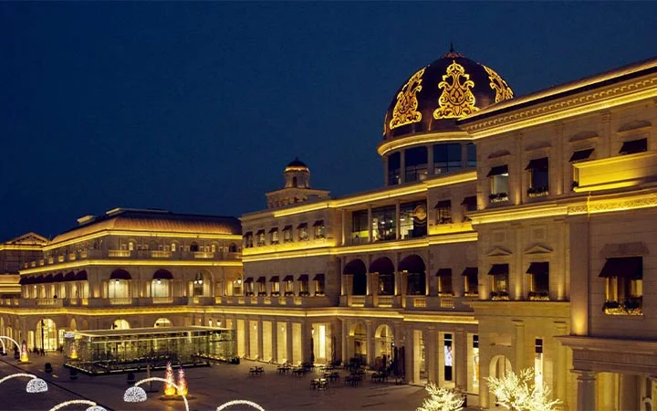 Elegant building with ornate architecture and a large, illuminated dome, glowing with golden lights at night. Reminiscent of Galeries Lafayette, its exterior features columns, arches, decorative details, and a courtyard visible in the foreground.