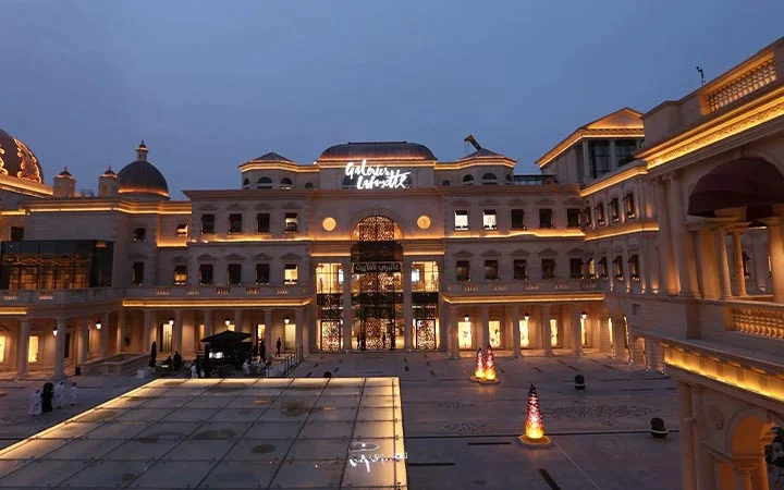 A grand shopping mall with elegant, palace-like architecture, warm glowing lights, and people strolling in the open courtyard at dusk. The illuminated sign Galeries Lafayette shines above the main entrance.