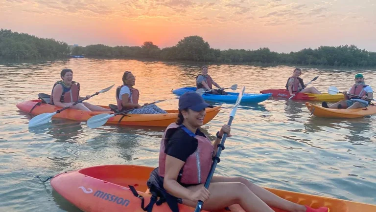 Six people in life vests paddle kayaks on calm water at sunset, surrounded by green trees. Smiling and relaxed, they enjoy the outdoor activity together—proof that Qatar is the Ultimate Destination for adventure and tranquility.