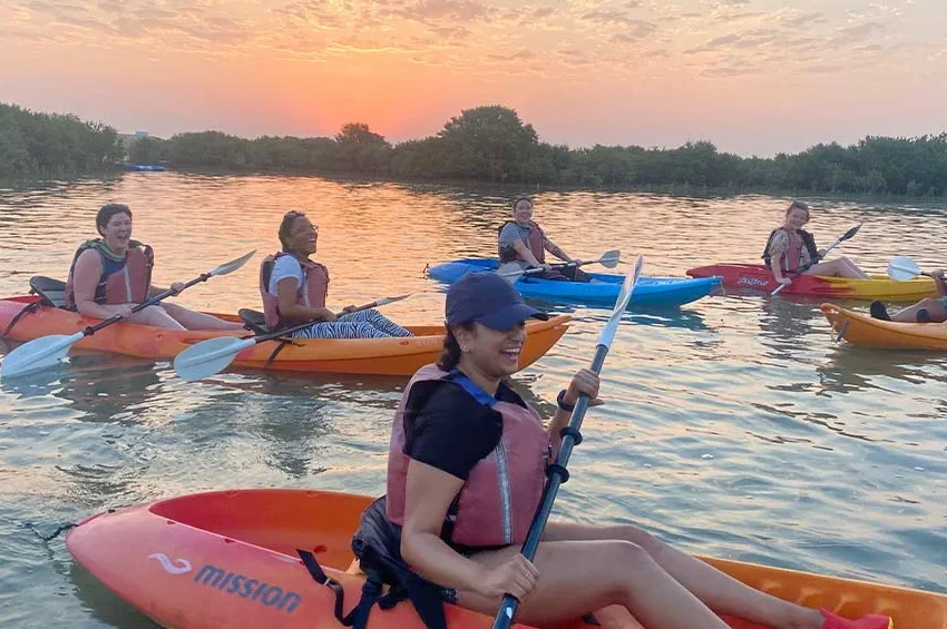 Six people in life vests paddle kayaks on calm water at sunset, surrounded by green trees. Smiling and relaxed, they enjoy the outdoor activity together—proof that Qatar is the Ultimate Destination for adventure and tranquility.