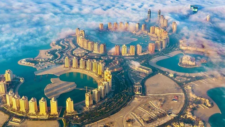Aerial view of The Pearl-Qatar, a man-made island in Doha popular for luxury travel Qatar, featuring clusters of high-rise buildings, turquoise lagoons, and sandy beaches, partly shrouded by low-hanging clouds.