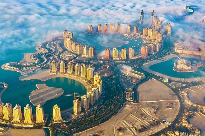 Aerial view of The Pearl-Qatar, a man-made island in Doha popular for luxury travel Qatar, featuring clusters of high-rise buildings, turquoise lagoons, and sandy beaches, partly shrouded by low-hanging clouds.