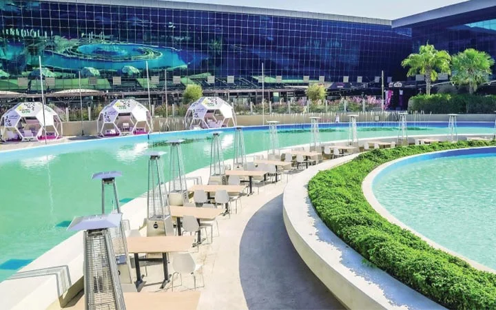 Outdoor dining tables are arranged beside a curved pool with water features at Lagoona Mall in Qatar. White cabanas line the pool’s edge, while a large glass building reflects palm trees and lush greenery in the background.