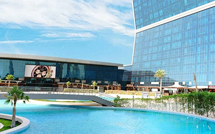 A modern, glass-fronted hotel building with a curved design stands beside a large swimming pool near Lagoona Mall in Qatar, surrounded by palm trees, a bridge, and a clear blue sky in the background.