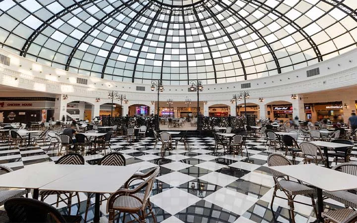 A spacious food court at Place Vendome Mall in Qatar features black and white checkered flooring, wicker chairs, and tables under a large glass dome ceiling. Several eateries line the curved walls, with a few people seated at the tables.