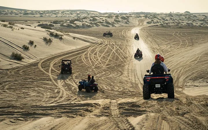 Several people enjoy a thrilling Desert Adventure as they ride ATVs and dune buggies across sandy, open desert terrain with tire tracks and scattered bushes under a hazy sky.