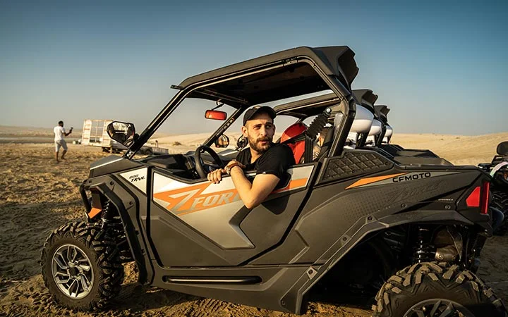 A man wearing a black cap and t-shirt sits in a black and orange off-road ATV, ready for a Desert Adventure, parked on sandy terrain with desert scenery and another person in the background.