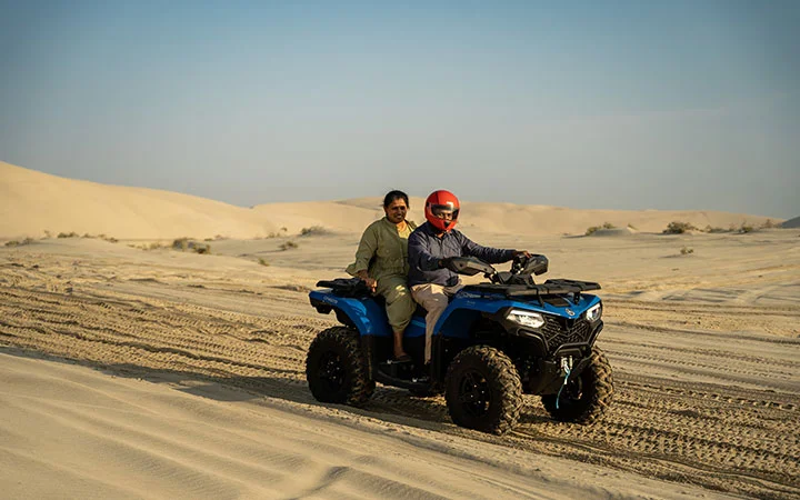 Two people enjoy a Desert Adventure, riding a blue quad bike across sandy dunes. The driver sports a red helmet and sunglasses, while the passenger sits behind. The clear sky and dry landscape add to this thrilling Quad Biking experience.