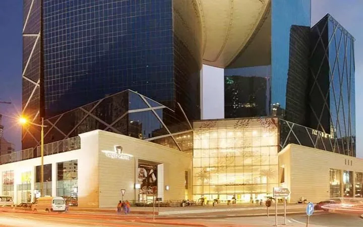Modern glass and steel skyscraper with a unique curved design, illuminated at dusk near The Gate Mall Qatar. The lower level features large glass windows and a brightly lit entrance, with cars and streetlights visible in the foreground.