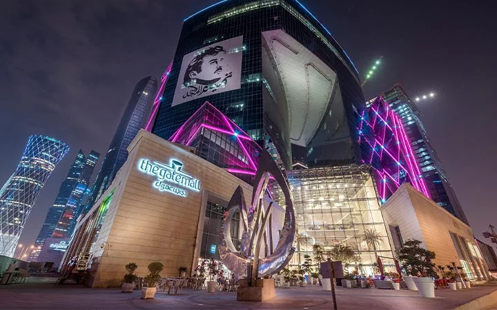 A nighttime view of The Gate Mall Qatar in Doha, featuring a modern glass building with vibrant pink and purple lights, a large outdoor sculpture, and illuminated signage. Skyscrapers and city lights shine in the background.