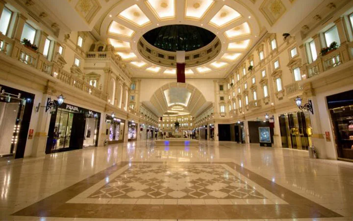 A spacious, elegant shopping mall interior inspired by Villaggio Mall Qatar, with bright lighting, high ceilings, marble floors, designer store fronts, and a large central dome. The mall appears empty and well-maintained.