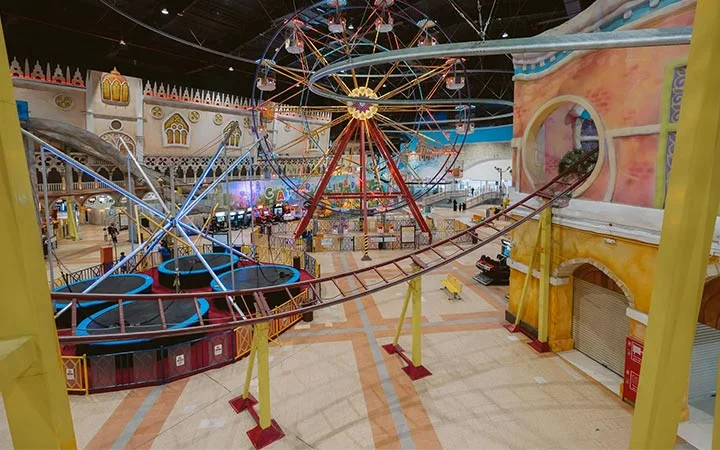 Experience an indoor amusement park at Villaggio Mall Qatar, featuring a Ferris wheel, roller coaster tracks winding around, arcade games, and colorful painted walls in a bright and spacious setting with playful, vibrant decor.