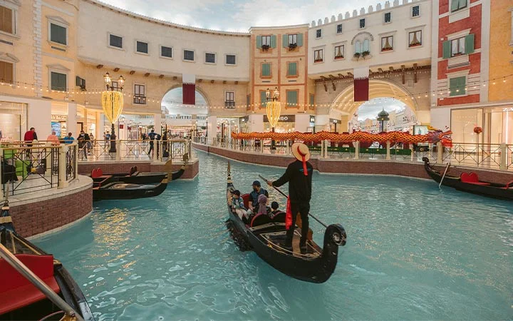 A gondolier steers a gondola with passengers along an indoor canal at Villaggio Mall Qatar, surrounded by European-style buildings and shops, beneath a sky-like ceiling in this lively shopping destination.